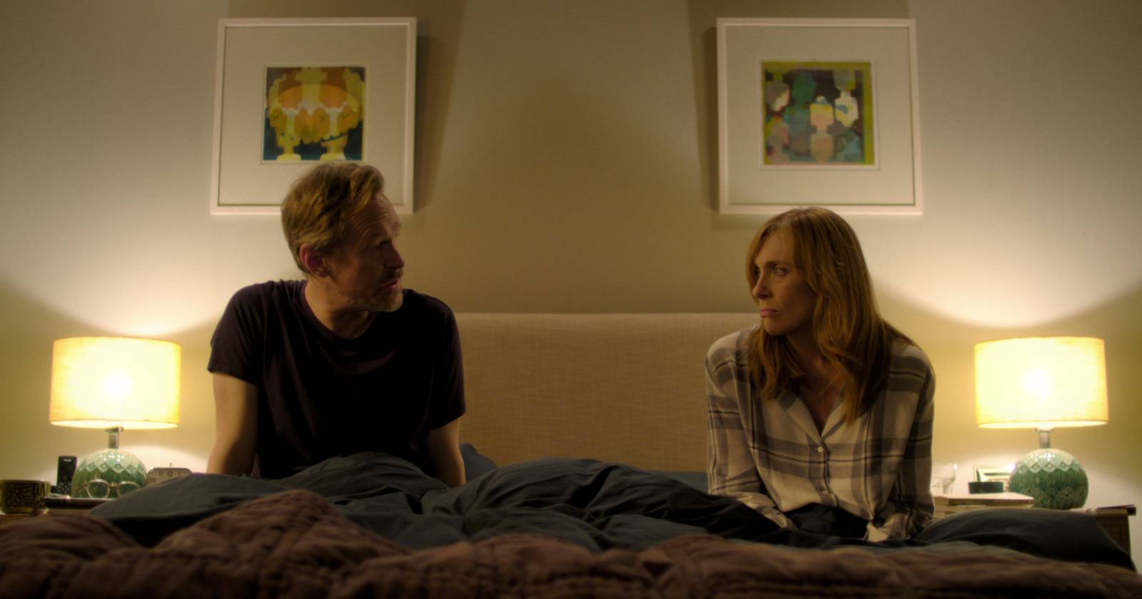 A la izquierda Steven Mackintosh como Alan, a la derecha Toni Collette como Joy. Ambos sentados en la cama conversando. En Wanderlust, serie original de Netflix.
