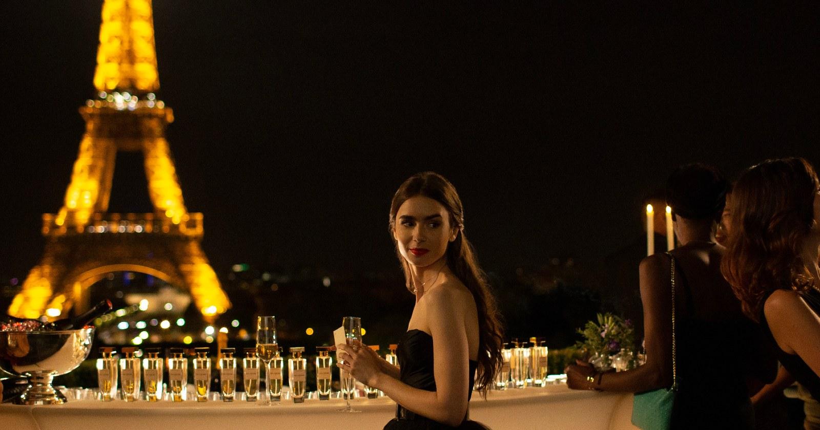 Lily Collins como Emily en París en una fiesta de gala nocturna con una copa de espumante en su mano y la Torre Eiffel con sus luces encendidas de fondo