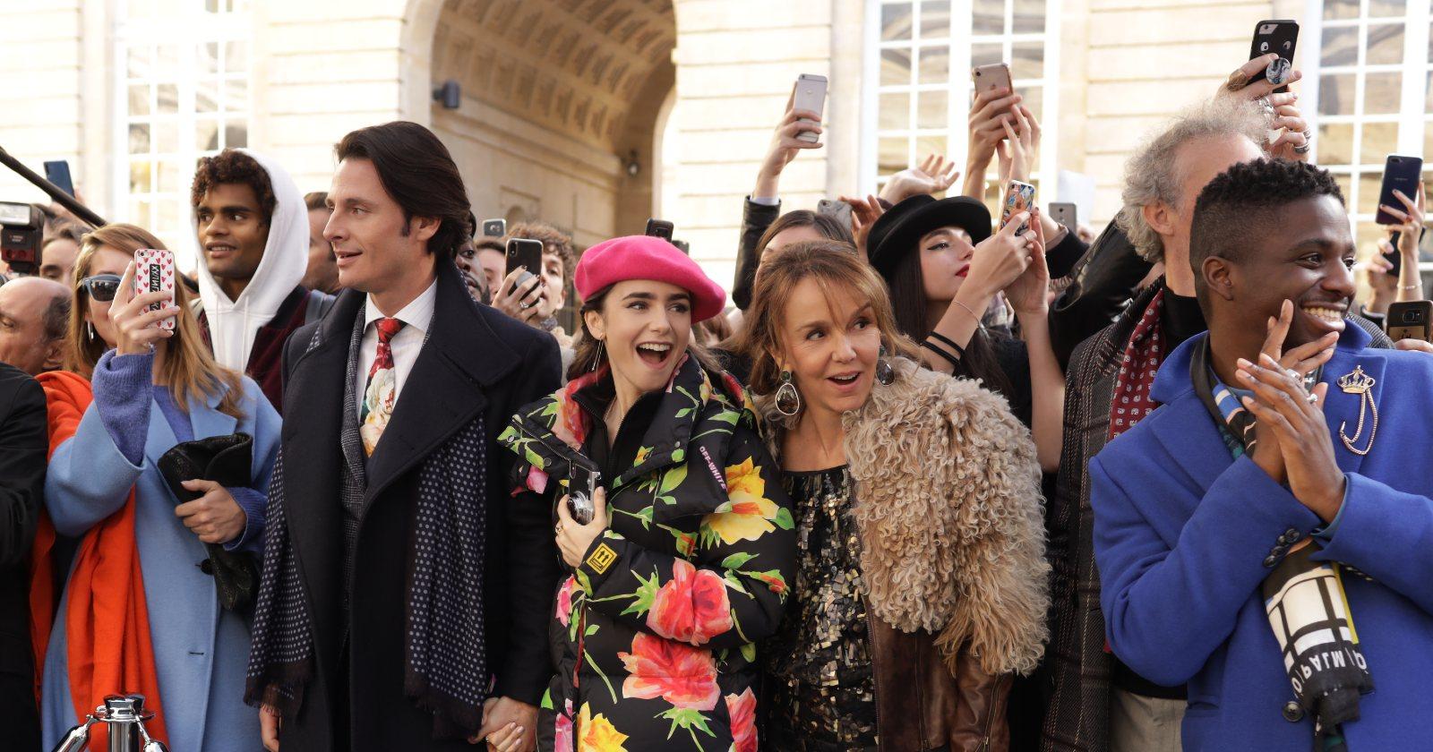 Emily en París en un desfile de modas en el exterior, en medio de una multitud tomando fotos con celulares, acompañada de sus colegas.