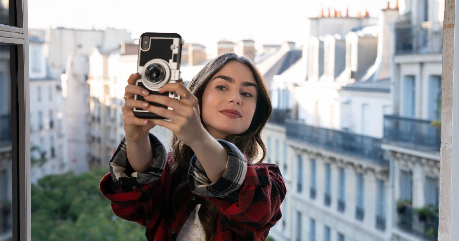 Emily en París en el balcón de su habitación Chambre de Bonne, tomándose una Selfie con los edificios blancos parisinos de fondo