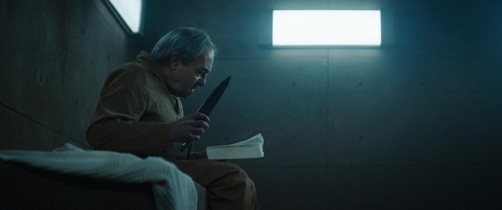 Zorion Eguileor como Trimagasi en El Hoyo sosteniendo un cuchillo en su mano mientras lee un libro