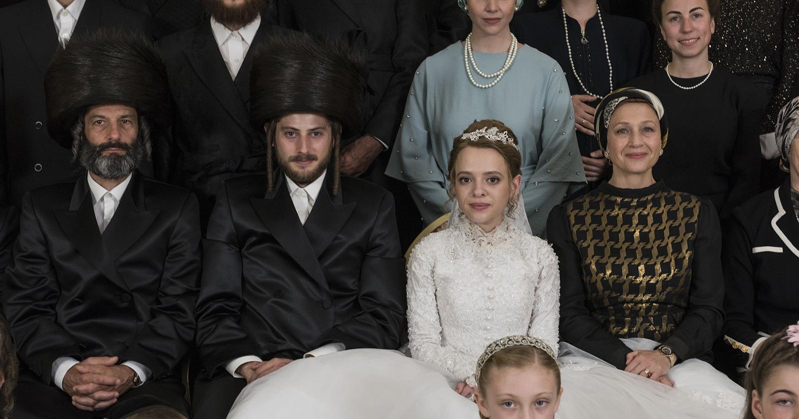 En la imagen vemos a Etsy, protagonista de la mini serie Poco Ortodoxa, en el día de su boda. Ella está sentada al medio de los miembros de su comunidad extremadamente religiosa. Esta fue una de las series 2020 más vistas de Netflix.