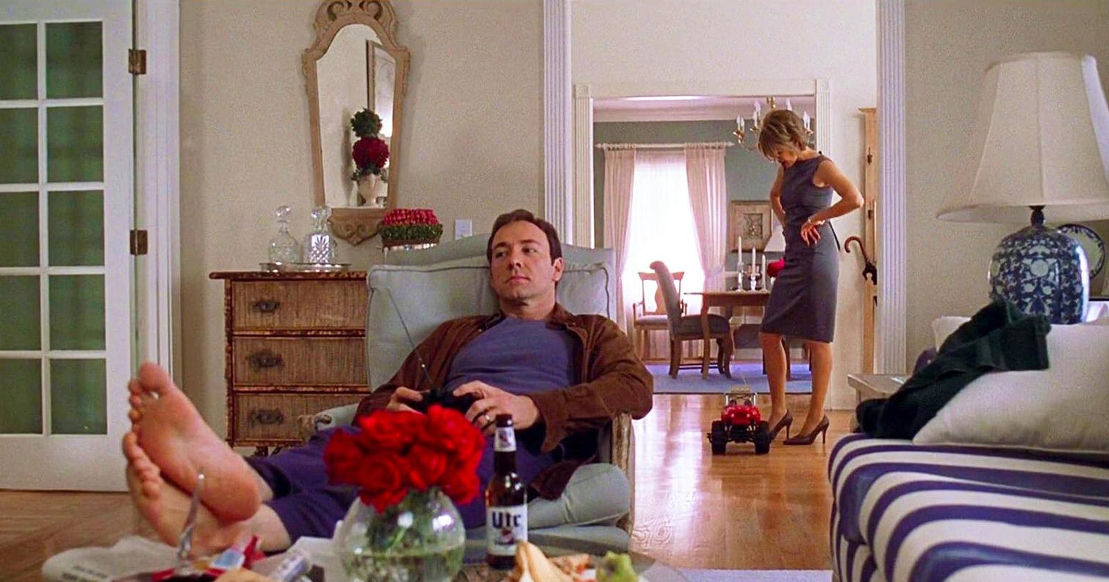 Lester (Kevin Spacey) tendido sobre un sillón. Detrás, Carolyn (Annette Bening) pasando la aspiradora. Escena de American Beauty.